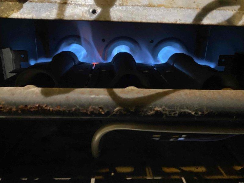 Performed seasonal tune-up on the gas furnace and air conditioning system. Inspected the furnace burner compartment, confirming stable blue flame across all burners, and noted surface rust and debris buildup on the burner compartment rail. Tested the outdoor condensing unit's electrical operation with a true RMS meter, recording 0.98 amps on the measured component, and verified the system was operating properly.