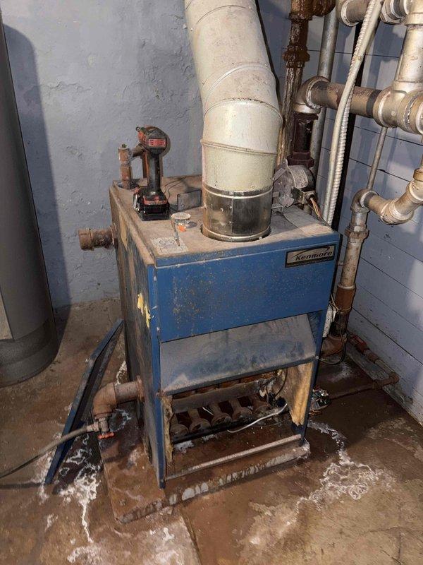 Responded to a no-heat call on an approximately 30-year-old Kenmore boiler and removed the lower access panel to inspect the burner tubes, combustion chamber, and control wiring. Diagnosis found significant corrosion at the piping fittings, standing water and residue pooled at the base of the unit, and deteriorated, tangled control wiring. The system's age and condition were documented with photos, and an estimate for repair or replacement will be provided.