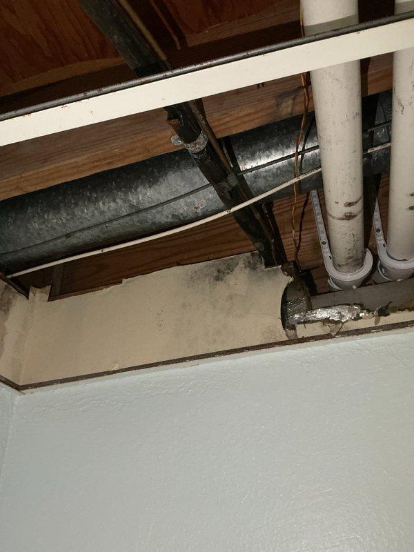 Inspected the 2-year-old A/C system in response to reported mold and moisture damage, and examined the exposed ceiling cavity where the supply ductwork and condensate drain piping run. Observed dark mold-like staining and moisture damage on the ceiling drywall directly beneath the galvanized duct and condensate drain line penetration, with torn duct wrap and disturbed insulation and patching at the opening. Documented the affected areas with photos and identified the condensate drain and duct insulation/penetration area as the probable moisture source.