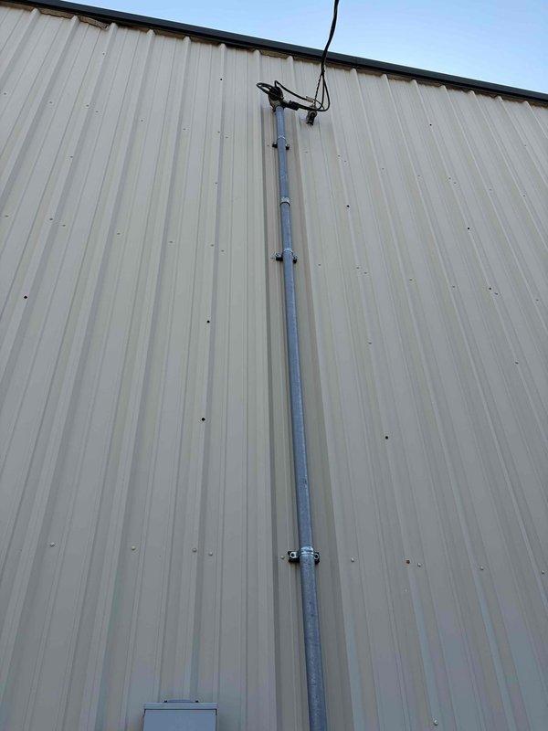 Completed site assessment for new 50-amp electrical service installation at commercial facility. The proposed service run measures approximately 65 feet from the existing customer service entrance disconnect (CSED) to the main building, requiring conduit routing across an exterior dirt lot area. Documented existing electrical infrastructure including weatherhead assembly, vertical conduit penetration through corrugated metal siding, and current panel configuration to determine material requirements and installation methodology for the new circuit.