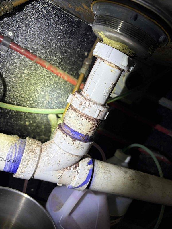 Inspected existing PVC drain line assembly for bar area sink and documented current plumbing configuration. The drain system consists of white PVC piping with multiple elbow fittings and connections showing age-related discoloration, mineral deposits, and rust staining around joints. Assessment revealed the need for replacement fixtures to properly complete the drain line installation. Photographed the existing pipe layout, including visible blue o-ring seals at connection points, chrome supply lines, and water filtration components for reference during fixture procurement and installation planning.