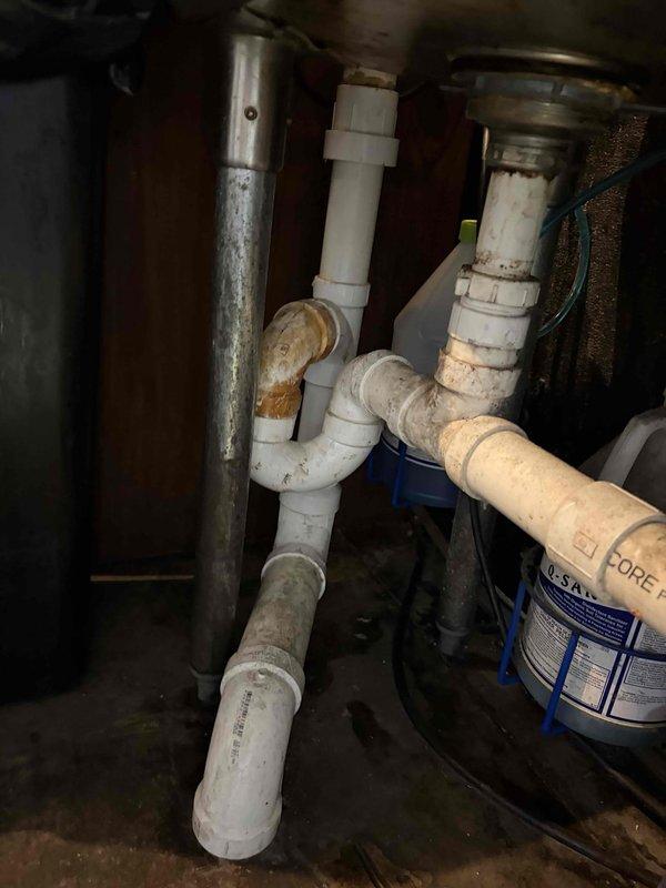 Inspected existing PVC drain line assembly for bar area sink and documented current plumbing configuration. The drain system consists of white PVC piping with multiple elbow fittings and connections showing age-related discoloration, mineral deposits, and rust staining around joints. Assessment revealed the need for replacement fixtures to properly complete the drain line installation. Photographed the existing pipe layout, including visible blue o-ring seals at connection points, chrome supply lines, and water filtration components for reference during fixture procurement and installation planning.