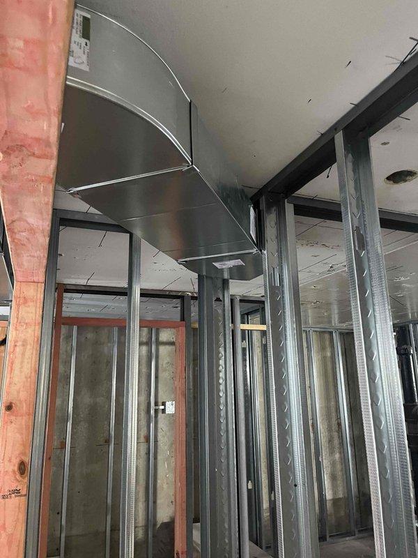 Continued commercial ductwork installation in interior construction phase. Installed large curved metal air duct runs along ceiling area, coordinating with existing metal stud framing. Work included integration with electrical pathways and preparation for upcoming phases around doorway openings and corridor sections.