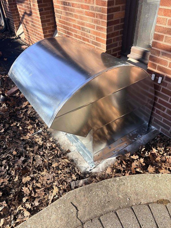 # Service Summary

Installed custom fabricated air intake hood over existing steel grate at boiler intake piping location to provide weather protection and improve system efficiency.

# Work Performed

Responded to commercial facility for installation of protective air intake hood assembly at boiler system intake point. Upon arrival, located existing steel grate where boiler intake piping penetrates building foundation at exterior wall. 

Fabricated and installed galvanized steel barrel-vault style hood enclosure to cover and protect the intake grate opening. Hood features rounded cylindrical top section transitioning to flat vertical side panels, providing complete weather protection while maintaining proper airflow to boiler intake system. Secured metal enclosure to brick exterior wall and foundation using appropriate fasteners for masonry application.

Ensured proper clearances around existing black conduit piping running vertically along brick wall. Verified hood installation does 