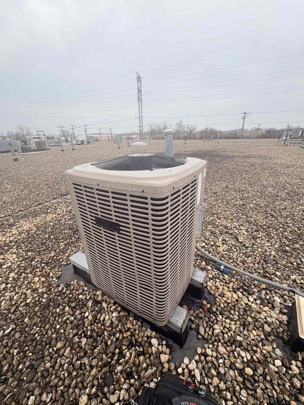 Responded to service call for non-operational air conditioning unit serving computer server room. Upon arrival, verified thermostat reading of 78°F and inspected Honeywell digital control mounted in mechanical room. Proceeded to rooftop to assess outdoor condensing unit condition. Performed visual inspection of rooftop-mounted condenser unit, noting equipment location on gravel surface with proper base pad installation. Documented system components including fan assembly, louvered cabinet, and surrounding HVAC infrastructure. Completed initial diagnostics within standard service timeframe and coordinated with office regarding additional diagnostic requirements and approval process for continued troubleshooting. Photographed thermostat display, wall-mounted controls, and exterior condensing unit for service documentation.