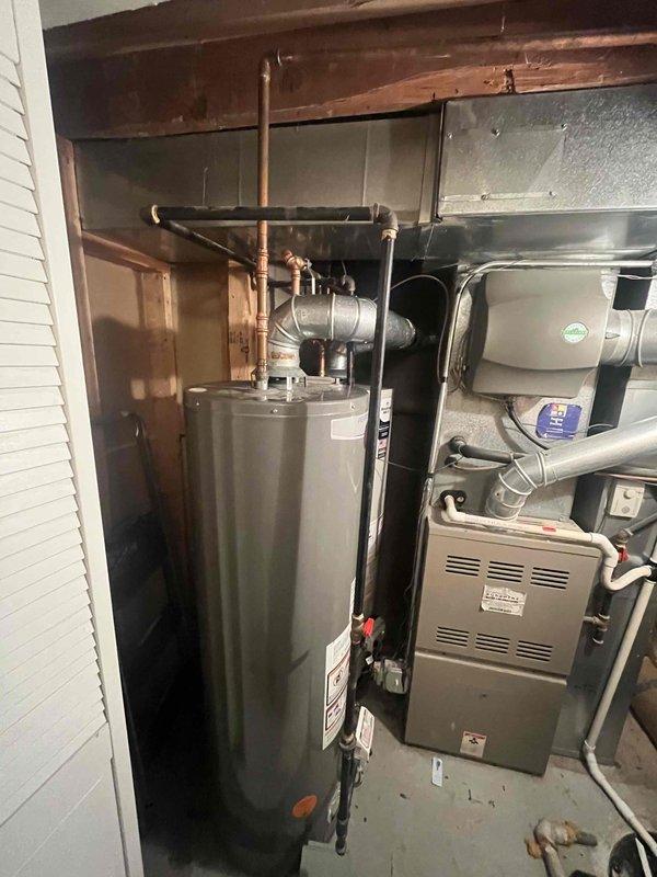 Completed tanked water heater installation in residential utility area with exposed beam construction. Installed new gray cylindrical water heater with proper copper piping connections, pressure relief valve, and all required fittings to code specifications. Relocated existing water heater to front position of unit as requested, requiring additional pipe fittings and labor to properly configure dual water heater setup. Both units now operational with appropriate clearances maintained alongside existing furnace and HVAC ductwork in mechanical room.