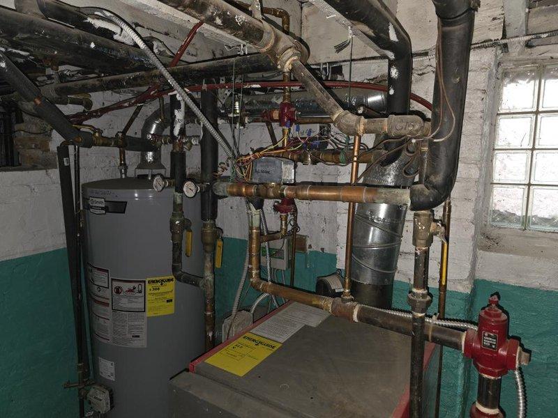 Conducted comprehensive assessment of existing HVAC and hydronic heating system for planned residential renovation project. Evaluated current basement mechanical room containing water heater, expansion tank, and complex piping network including copper, black insulated, and color-coded distribution lines with multiple valves and gauges. Documented existing two-tone painted mechanical space with overhead piping infrastructure and window access. Reviewed architectural plans specifying retention of select radiators while replacing others with baseboard units. Assessed feasibility of installing new combination hot water/boiler system in basement to serve primary heating zones. Evaluated existing R410A outdoor compressor unit for compatibility and potential reuse with planned Space-Pak high-velocity air conditioning system to be installed in attic location near current equipment. Confirmed separate inline water heater specification for two new attic bathroom additions independent of main hea