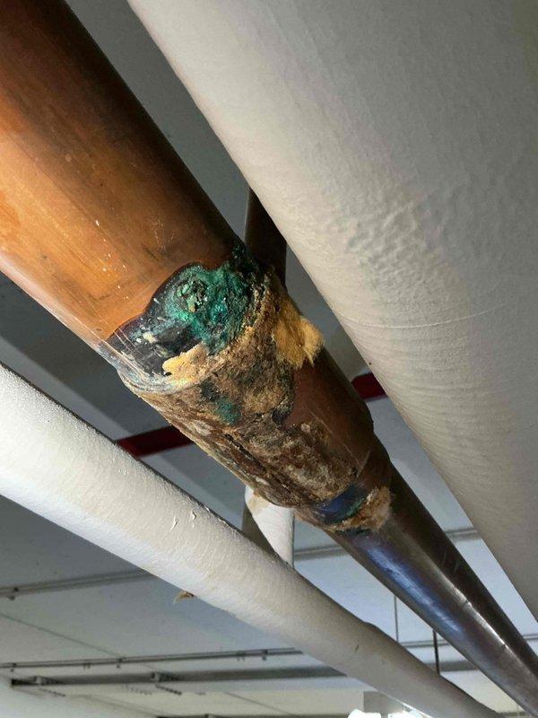Responded to emergency water leak call in commercial building garage area affecting heating system piping leading to boiler room. Upon arrival, coordinated building access and conducted thorough inspection of the overhead piping infrastructure. Located source of leak at corroded copper pipe joint showing extensive green patina, verdigris buildup, and surface deterioration from prolonged moisture exposure. Documented pipe measurements and corrosion damage with photographs for repair assessment. The affected heating pipe joint exhibited severe oxidation and mineral deposits requiring replacement rather than simple repair due to extent of deterioration.