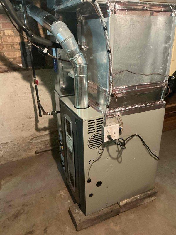 Completed installation of residential furnace system in basement location. Unit was properly positioned on wooden base over concrete floor, with metallic ductwork connected and secured using foil tape, electrical connections made to outlet and control box, and all wiring routed through black flexible conduit. System installation verified and remaining balance collected upon completion.