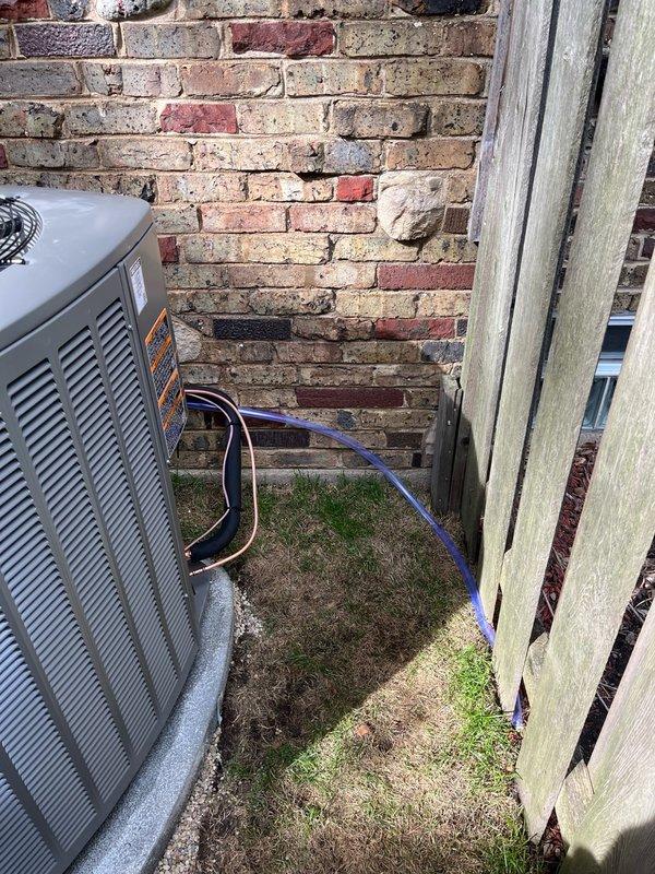 Continued and completed high velocity HVAC system installation at residential property. Installed Lennox outdoor condensing unit on concrete pad in side yard between brick exterior wall and privacy fence, connected refrigerant lines with proper insulation (black foam and blue covering), and routed lines along fence structure. System positioned for optimal airflow and serviceability in confined space, with all refrigerant piping secured and electrical connections completed per manufacturer specifications.