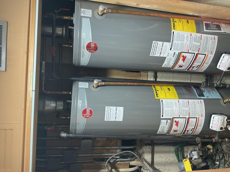 Completed installation of two 40-gallon tanked water heaters, replacing the existing two 50-gallon units. Both water heaters were positioned side by side in the utility area, with all copper supply lines, brass fittings, pressure relief valves, and drainage connections properly installed and secured according to code requirements.