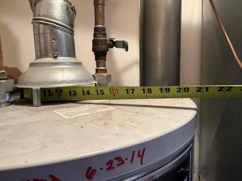 Responded to urgent water leak from 12-year-old residential water heater affecting adjacent bedroom area. Upon inspection, documented unit dimensions and assessed leak source at plumbing connections and valve fittings on top of unit. Measured clearances and evaluated water damage extent at doorway threshold where moisture had penetrated flooring, documenting approximately 5-7 inches of affected area between door frame and wall surface.
