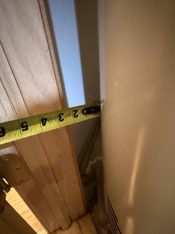 Responded to urgent water leak from 12-year-old residential water heater affecting adjacent bedroom area. Upon inspection, documented unit dimensions and assessed leak source at plumbing connections and valve fittings on top of unit. Measured clearances and evaluated water damage extent at doorway threshold where moisture had penetrated flooring, documenting approximately 5-7 inches of affected area between door frame and wall surface.