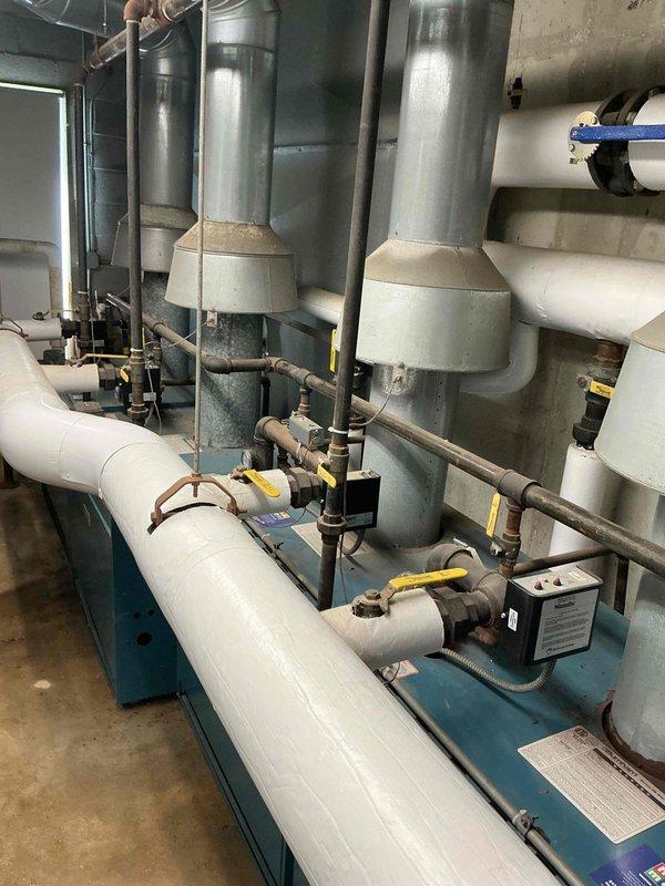 Responded to emergency water leak at commercial boiler system. Upon arrival, found that during pressure relief valve testing, the rear section of the boiler began discharging water rapidly from the manifold area. Inspected the brass manifold assembly and associated piping connections within the boiler cabinet and identified compromised seal integrity at multiple connection points showing corrosion and age-related deterioration. Documented equipment condition including the multi-unit boiler installation with vertical flues, insulated distribution piping, and control valving. Photographed the internal manifold system showing discoloration and buildup on brass pipes, as well as the gas valve assembly and associated copper tubing connections. System was secured to prevent further water discharge and customer was advised of findings for repair authorization.
