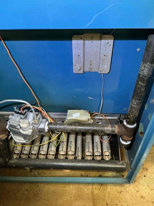 Responded to emergency water leak at commercial boiler system. Upon arrival, found that during pressure relief valve testing, the rear section of the boiler began discharging water rapidly from the manifold area. Inspected the brass manifold assembly and associated piping connections within the boiler cabinet and identified compromised seal integrity at multiple connection points showing corrosion and age-related deterioration. Documented equipment condition including the multi-unit boiler installation with vertical flues, insulated distribution piping, and control valving. Photographed the internal manifold system showing discoloration and buildup on brass pipes, as well as the gas valve assembly and associated copper tubing connections. System was secured to prevent further water discharge and customer was advised of findings for repair authorization.