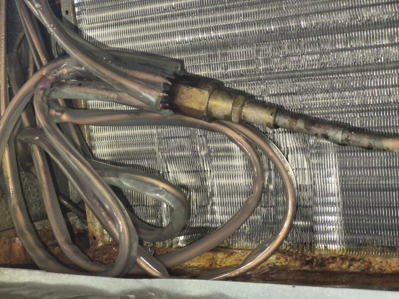 Responded to service call for commercial ice cream shop experiencing inadequate cooling performance. Upon inspection, found evaporator coil with significant corrosion and oxidation on copper refrigerant lines, indicating system age and possible refrigerant line degradation. Observed corroded brass fittings at connection points and discolored metal fins with dirt accumulation affecting heat transfer efficiency. Exterior condensate drain system showed proper drainage function with weathered galvanized drain pan and PVC discharge line intact. Documented equipment condition showing normal wear patterns consistent with extended service life and recommended monitoring for potential refrigerant leaks due to copper line deterioration.