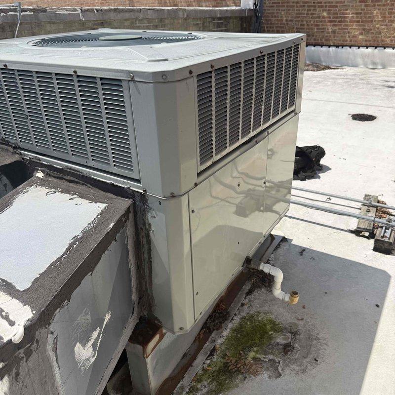 Responded to commercial rooftop unit (RTU) after 12 PM for scheduled maintenance agreement service per customer contact person. Upon arrival, accessed RTU mounted on rooftop curb and performed comprehensive system inspection. Unit found operational with condenser fan assembly functioning properly. Documented equipment condition with photographs showing beige packaged rooftop unit with horizontal louver ventilation panels. Noted rooftop membrane showing signs of weathering, algae growth, and standing water accumulation on surrounding roof surface. HVAC unit mounting platform exhibited deterioration with peeling coating and dark staining at base where platform meets roof membrane. Completed routine maintenance checks and verified proper airflow through louver vents and fan grille assembly. System operating within normal parameters at time of service completion.