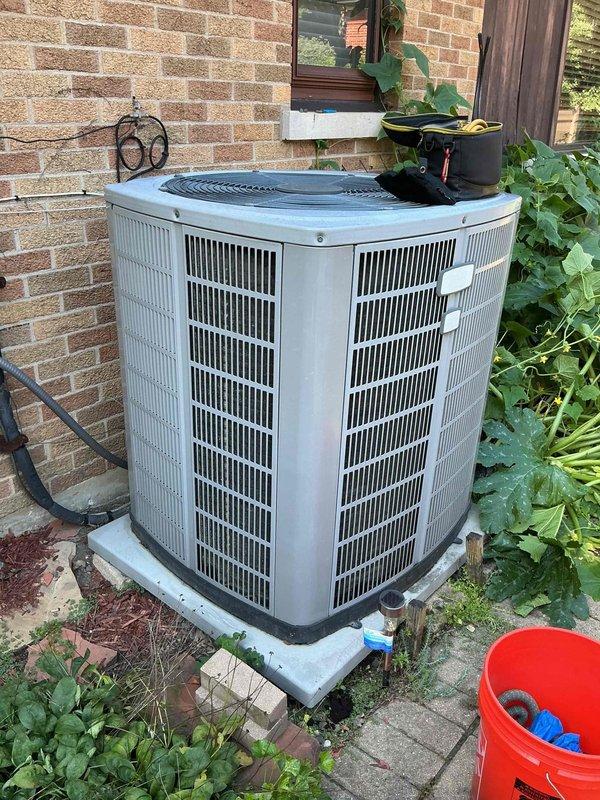 Responded to no cooling complaint on aging residential air conditioning system. Upon arrival, located outdoor condenser unit positioned against exterior brick wall on concrete pad. Performed visual inspection of Rheem HVAC system including indoor furnace unit and outdoor condensing unit. Verified thermostat settings and confirmed unit was set to cooling mode. Checked electrical disconnect and circuit breakers for proper operation. Inspected refrigerant line set entering building and examined electrical conduit connections. Evaluated condensate pump operation at indoor unit. Assessed overall system condition noting advanced age of equipment. Documented equipment information and captured photographic evidence of both indoor and outdoor components for service records.