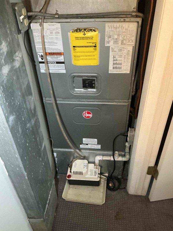 Responded to no cooling complaint on aging residential air conditioning system. Upon arrival, located outdoor condenser unit positioned against exterior brick wall on concrete pad. Performed visual inspection of Rheem HVAC system including indoor furnace unit and outdoor condensing unit. Verified thermostat settings and confirmed unit was set to cooling mode. Checked electrical disconnect and circuit breakers for proper operation. Inspected refrigerant line set entering building and examined electrical conduit connections. Evaluated condensate pump operation at indoor unit. Assessed overall system condition noting advanced age of equipment. Documented equipment information and captured photographic evidence of both indoor and outdoor components for service records.