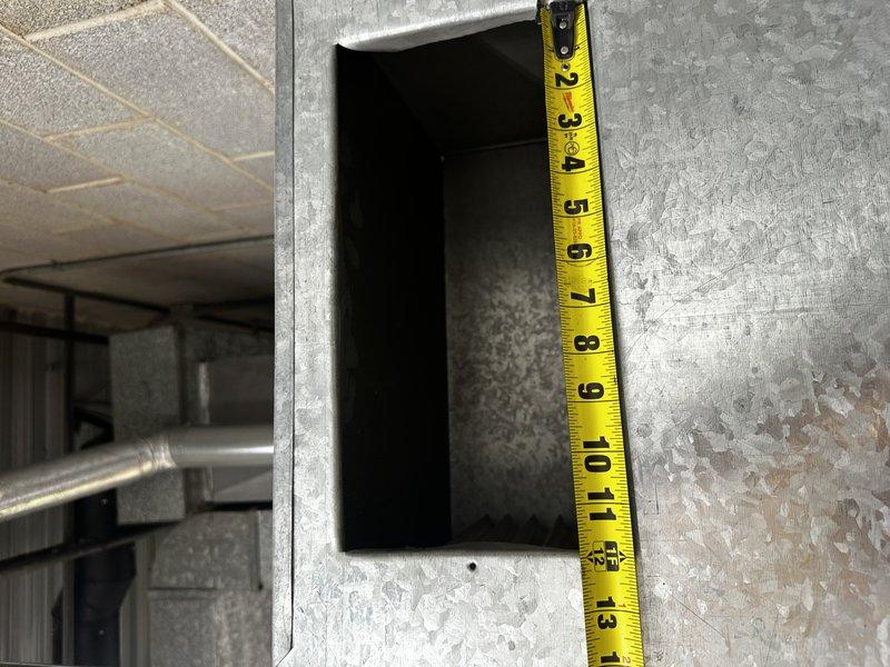 Performed field measurements of return air opening and ductwork on warehouse furnace system to provide accurate dimensions for commercial HVAC estimate. Documented return opening height at 13 inches and captured measurements of existing rectangular metal duct connections on the air handler unit. Photographed overhead HVAC installation including large rectangular sheet metal ductwork, air handler with blower compartment, and associated black piping connections mounted near drop ceiling for estimation and planning purposes.
