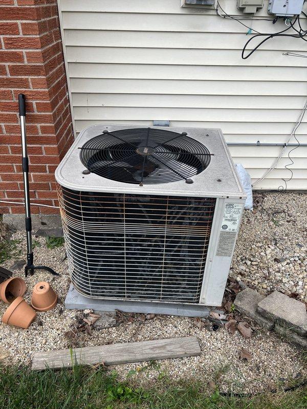 Conducted comprehensive site evaluation for complete HVAC system replacement including furnace and air conditioning unit. Assessed existing operational equipment with furnace located in basement and outdoor condenser unit positioned on gravel pad against exterior wall near brick chimney. Documented current system configuration, electrical connections, and installation conditions. Photographed outdoor condenser unit showing beige/tan metal cabinet with horizontal louvers, top-mounted circular fan assembly, electrical disconnect box, and meter location. Prepared replacement estimate for full heating and cooling system upgrade.