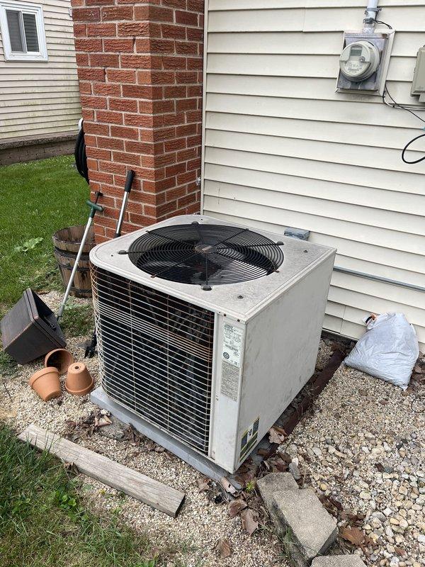 Conducted comprehensive site evaluation for complete HVAC system replacement including furnace and air conditioning unit. Assessed existing operational equipment with furnace located in basement and outdoor condenser unit positioned on gravel pad against exterior wall near brick chimney. Documented current system configuration, electrical connections, and installation conditions. Photographed outdoor condenser unit showing beige/tan metal cabinet with horizontal louvers, top-mounted circular fan assembly, electrical disconnect box, and meter location. Prepared replacement estimate for full heating and cooling system upgrade.