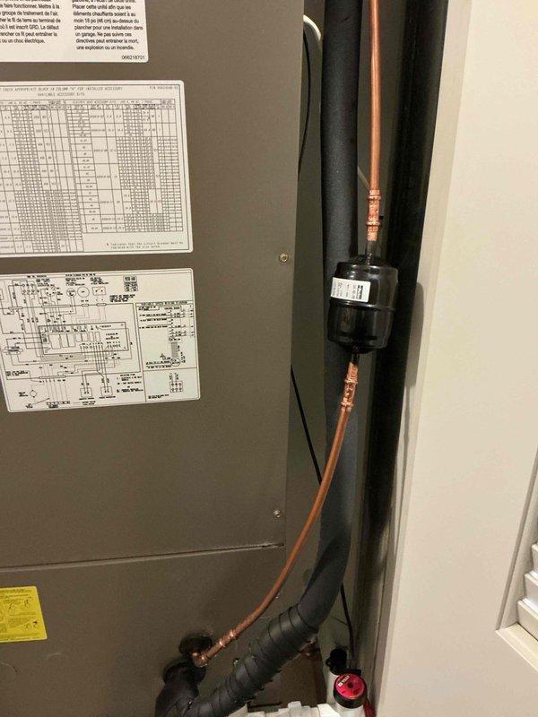 Completed warranty installation of suction line filter-drier on recently installed HVAC system where component was initially omitted. Installed black cylindrical filter-drier inline with copper refrigerant lines using proper soldering techniques and refrigeration line installation methods. Component was properly mounted vertically along wall adjacent to air handler unit with appropriate copper connections on both ends. All refrigerant lines, fittings, and solder joints were verified for proper installation and system integrity.