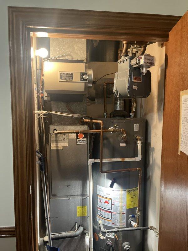 Completed on-site evaluation of existing residential HVAC and water heating equipment for replacement estimate. Documented current mechanical closet configuration containing gray Modello furnace unit, cylindrical water heater with specification labels, ceiling-mounted humidifier system, and associated copper plumbing, gas lines, and electrical connections. Assessed equipment accessibility, spatial constraints, and existing utility connections including flexible gas line routing and copper pipe configurations. Photographed current installation layout showing vertical arrangement of furnace, water heater, and humidifier components within confined closet space with wood trim finish. Evaluated connection points for water supply lines, shut-off valves, ductwork, and electrical conduit to determine replacement requirements and compatibility with new equipment specifications.