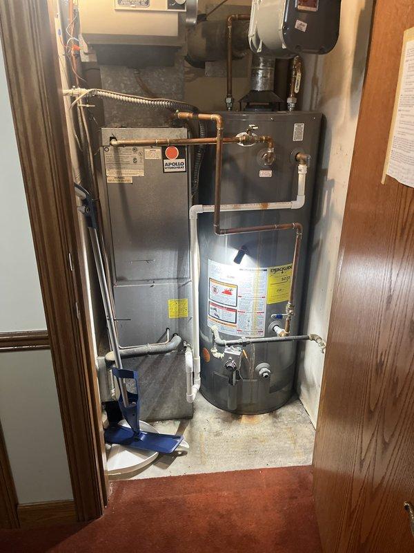 Completed on-site evaluation of existing residential HVAC and water heating equipment for replacement estimate. Documented current mechanical closet configuration containing gray Modello furnace unit, cylindrical water heater with specification labels, ceiling-mounted humidifier system, and associated copper plumbing, gas lines, and electrical connections. Assessed equipment accessibility, spatial constraints, and existing utility connections including flexible gas line routing and copper pipe configurations. Photographed current installation layout showing vertical arrangement of furnace, water heater, and humidifier components within confined closet space with wood trim finish. Evaluated connection points for water supply lines, shut-off valves, ductwork, and electrical conduit to determine replacement requirements and compatibility with new equipment specifications.