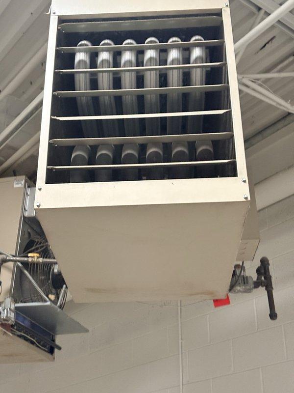 Performed scheduled preventive maintenance on a ceiling-suspended gas-fired unit heater, inspecting the cabinet, louvered grille, heat exchanger, motor and fan assembly, flexible gas connector, and black iron gas piping with shutoff valve; unit appeared intact with only minor surface dirt and light staining at the lower panel, and no frost or significant rust observed. Verified wall-mounted Honeywell thermostat operation in heat mode with a 73° setpoint and "Heat On" call displayed, noting dust and grime buildup on the thermostat housing. Documented equipment and filter information for the maintenance checklist; no filter list changes were identified.