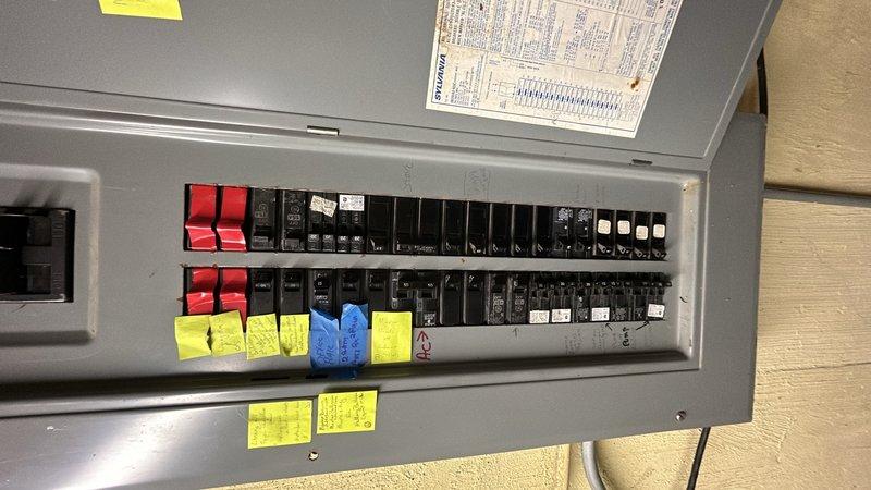 Performed comprehensive assessment of existing electrical panel for replacement quote. Documented current panel configuration showing multiple circuit breakers with several red-handled breakers indicating potential Federal Pacific or similar older equipment requiring replacement. Photographed panel interior revealing circuit layout, existing labeling system with yellow notes, and overall condition of breaker arrangement. Also documented related electrical components including wall-mounted thermostat or control device to ensure compatibility considerations for new panel installation.