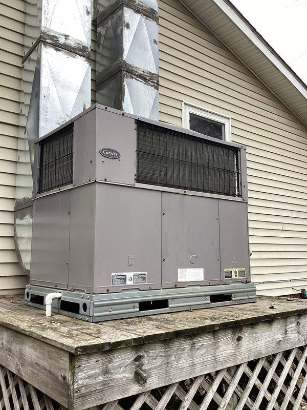 Responded to emergency heat call for older Carrier system. Inspected outdoor unit which was mounted on aging wooden platform with visible weathering. Found thermostat in emergency heat mode displaying 70° indoor temperature while set to 69°. Diagnosed failed heat pump operation causing system to rely on auxiliary heat. Restored primary heating function by replacing defective contactor and cleaning restricted condenser coil, restoring efficient operation.