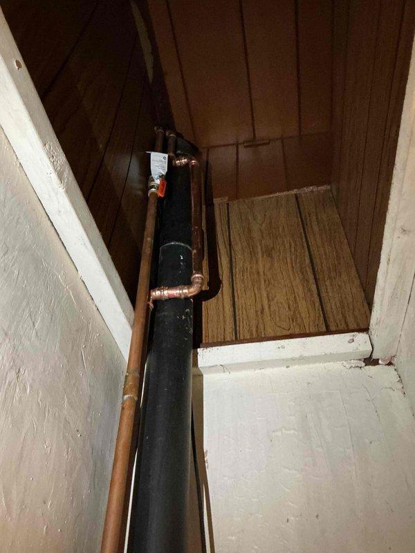Installed vertical black insulated water pipe with copper T-connection in utility closet corner. Completed soldering of copper pipe joints and attached identification tag at junction point. All connections were properly fitted and secured to both drywall and wood-paneled surfaces.