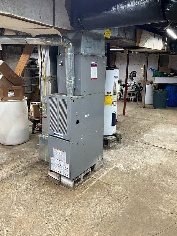 Performed inspection of 23-24 year old Tempstar furnace installed in basement utility area. Customer seeking replacement due to age of unit. Current setup includes furnace mounted on concrete platform with silver ductwork extending upward, positioned adjacent to water heater. Documented existing installation configuration to prepare appropriate replacement proposal.