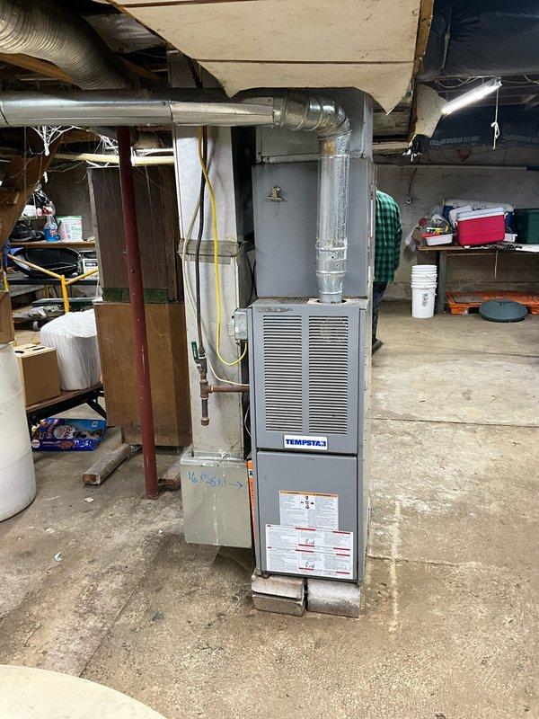 Performed inspection of 23-24 year old Tempstar furnace installed in basement utility area. Customer seeking replacement due to age of unit. Current setup includes furnace mounted on concrete platform with silver ductwork extending upward, positioned adjacent to water heater. Documented existing installation configuration to prepare appropriate replacement proposal.