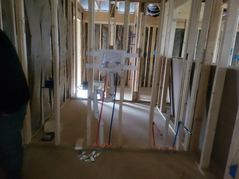 Completed rough-in plumbing installation in commercial construction site, installing PEX water supply lines (hot and red, cold and blue) through floor penetrations and securing them to wooden framing. Installed white PVC drainage pipes vertically through framing structure, ensuring proper alignment and support prior to insulation and drywall phases.