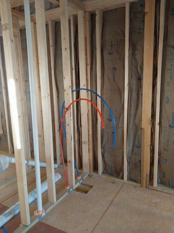 Completed rough-in plumbing installation in commercial construction site, installing PEX water supply lines (hot and red, cold and blue) through floor penetrations and securing them to wooden framing. Installed white PVC drainage pipes vertically through framing structure, ensuring proper alignment and support prior to insulation and drywall phases.