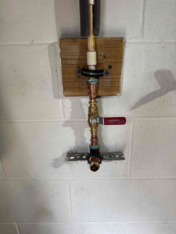 Inspected and repaired a leaking faucet installation in garage, which consisted of copper and brass pipe assembly mounted on a wooden board against concrete block wall. Observed water staining on mounting board indicating previous leakage, replaced deteriorated components, and verified proper operation of the brass shut-off valve with the red handle tag to ensure complete resolution of the leak issue.