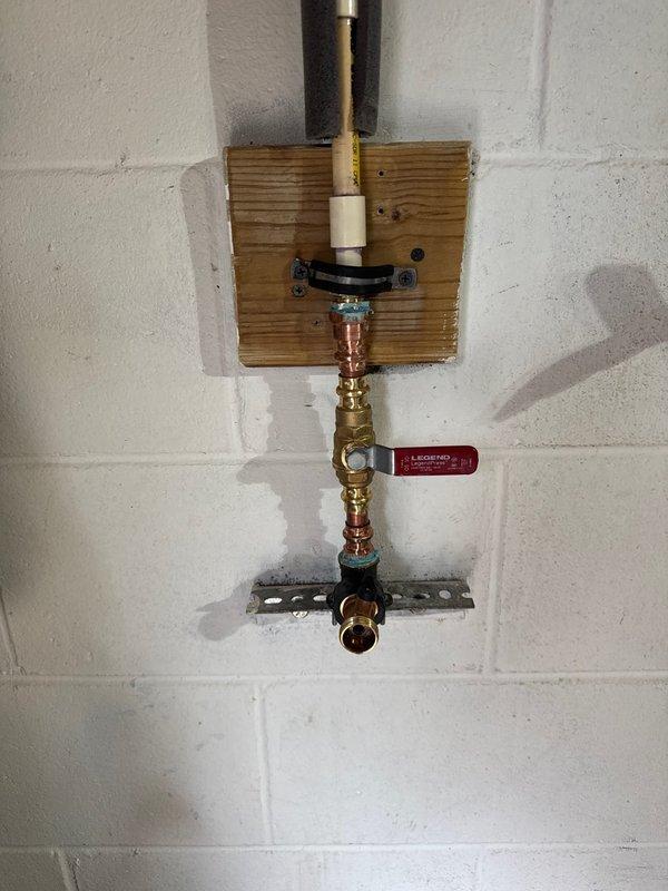 Inspected and repaired a leaking faucet installation in garage, which consisted of copper and brass pipe assembly mounted on a wooden board against concrete block wall. Observed water staining on mounting board indicating previous leakage, replaced deteriorated components, and verified proper operation of the brass shut-off valve with the red handle tag to ensure complete resolution of the leak issue.