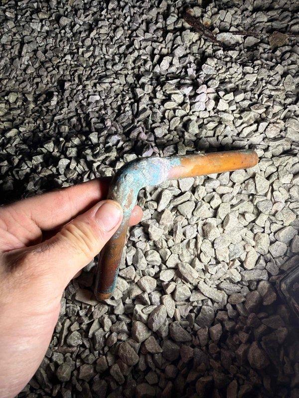 Diagnosed humming noise under bathroom toilet as deteriorated copper supply piping. Located corroded copper elbow fitting showing significant oxidation and patina formation. Replaced damaged section of copper plumbing and installed new shutoff valve to resolve noise issue. Verified proper water flow and absence of leaks after repair.