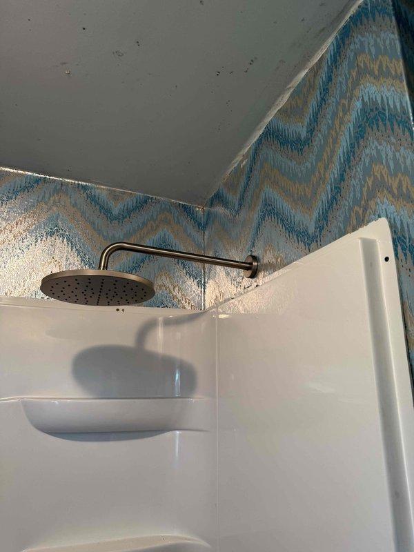 Completed installation of walk-in shower unit in RV bathroom, featuring white molded fiberglass construction with integrated shelving recesses. Mounted chrome shower head assembly on curved arm extension and installed decorative chevron-patterned tile work on upper walls in blue, teal, and beige tones to complement existing bathroom design.