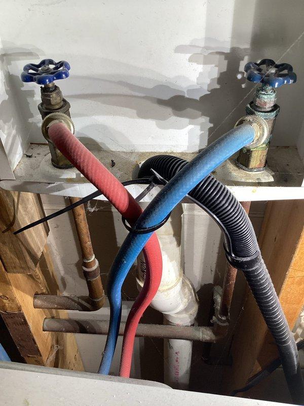 Completed monthly membership plumbing inspection of residential property. Inspected washing machine supply valves and connections, noting aged brass valves with blue handles, foam-insulated hot and cold supply lines, and corrugated flexible drain hose—all connections secure with no active leaks detected. Examined under-sink plumbing including PVC drain assembly and braided supply line with shutoff valve, confirming proper operation and drainage with no visible leaks or immediate concerns requiring attention.