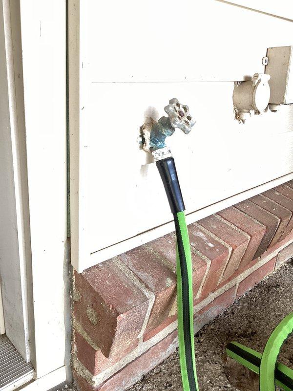 Responded to service call for exterior hose bib with garden hose frozen to connection point. Upon arrival, inspected chrome-bodied spigot mounted on white wood siding exterior wall above brick foundation ledge. Assessed condition of valve assembly with blue-gray handle mechanism and green garden hose attachment showing freeze damage at coupling point. Successfully removed frozen hose from spigot connection, verified valve operation and tested for leaks. Confirmed spigot functionality with no replacement required at this time.