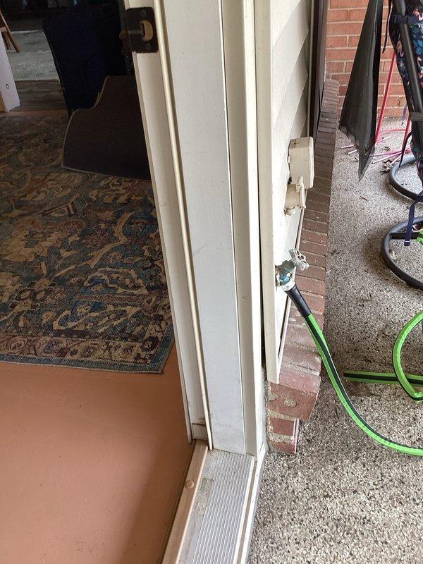 Responded to service call for exterior hose bib with garden hose frozen to connection point. Upon arrival, inspected chrome-bodied spigot mounted on white wood siding exterior wall above brick foundation ledge. Assessed condition of valve assembly with blue-gray handle mechanism and green garden hose attachment showing freeze damage at coupling point. Successfully removed frozen hose from spigot connection, verified valve operation and tested for leaks. Confirmed spigot functionality with no replacement required at this time.