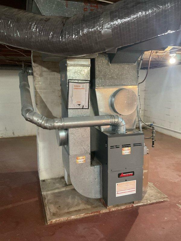 Conducted on-site assessment for complete HVAC system replacement at residential property. Evaluated existing furnace installation in basement utility area, documenting current equipment configuration including ductwork layout, return air plenum, and supply trunk lines. Photographed existing dark gray furnace unit with connected galvanized metal supply ductwork and flexible insulated ducting running along ceiling joists. Noted wall-mounted air handler or filter cabinet positioned adjacent to main heating unit. Assessed concrete floor mounting pad condition and existing duct connections for compatibility with new equipment installation. Documented current system specifications including manufacturer labels and equipment ratings visible on existing units. Evaluated ductwork condition, noting accumulated debris at seams and connections that will require cleaning during replacement. Measured clearances and access points for equipment removal and new unit installation. Prepared comprehensiv