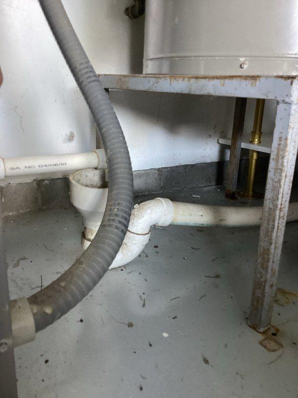 Completed monthly recurring plumbing inspection of residential water heater and associated plumbing systems. Documented condition of Reliance water heater unit, copper supply lines showing surface oxidation, flexible connector assembly, and PVC drainage piping in utility cabinet with visible age-related discoloration and rust staining on enclosure frame.