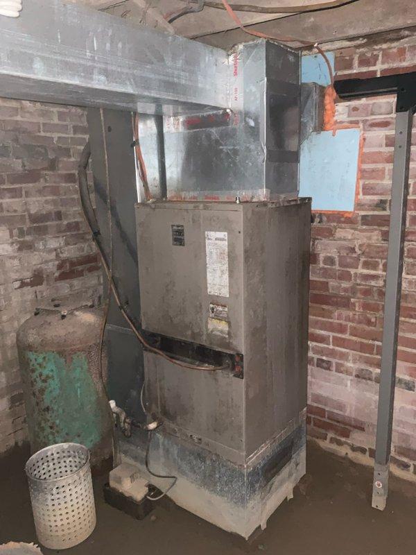 Conducted on-site assessment of non-operational HVAC system at rental property to provide replacement estimate. Inspected aging furnace unit from the 1980s-1990s located in basement utility area with exposed brick walls, noting significant dust accumulation, surface wear, and deterioration of surrounding components including corroded cylindrical equipment and discolored electrical panel with Square D breakers showing heavy grime buildup. Documented existing equipment condition and ductwork configuration through photographs for replacement system specifications.