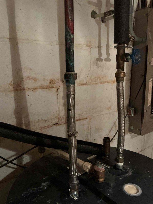Responded to water leak investigation initially suspected to be from AC unit. Upon inspection, determined leak source was from piping connected to the top of the water heater. Inspected flexible braided metal supply lines, valve connections, and associated plumbing assembly in utility area. Documented plumbing configuration including stainless steel braided hoses, copper piping, valve with blue handle, and black iron pipes. Noted evidence of previous water damage and staining on concrete block walls. Confirmed best contact number and photographed affected plumbing components for documentation purposes.
