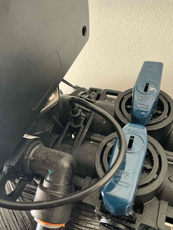 Responded to service call for leaking water softener that was recently activated by new homeowner. Upon inspection, identified active leak from softener system and assessed condition of unit and connections. Documented equipment condition with photographs showing water softener components, bypass valve assembly with blue control switches in OFF position, and associated plumbing connections including black flexible hoses and fittings that exhibited signs of age and wear.