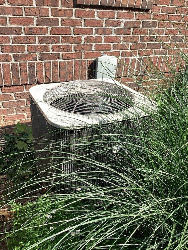 # HVAC Service Call - No Cooling Complaint

Responded to no cooling complaint on residential air conditioning system over 10 years old. Upon arrival, found outdoor condensing unit heavily obstructed by overgrown vegetation and debris accumulation in mulch bed surrounding equipment. Unit showed signs of age-related deterioration including oxidation on refrigerant lines and general weathering of components.

Cleared dense grass and weeds growing around condenser cabinet that were blocking airflow to unit and restricting proper ventilation through side panels. Removed leaves, organic debris, and accumulated material from around equipment base and refrigerant line penetrations. 

Inspected condenser coil fins which displayed discoloration and contamination buildup. Assessed refrigerant line insulation condition - noted degradation and exposure on suction line. Examined electrical disconnect and service panel mounted on exterior wall for proper operation.

Performed visual inspection of all
